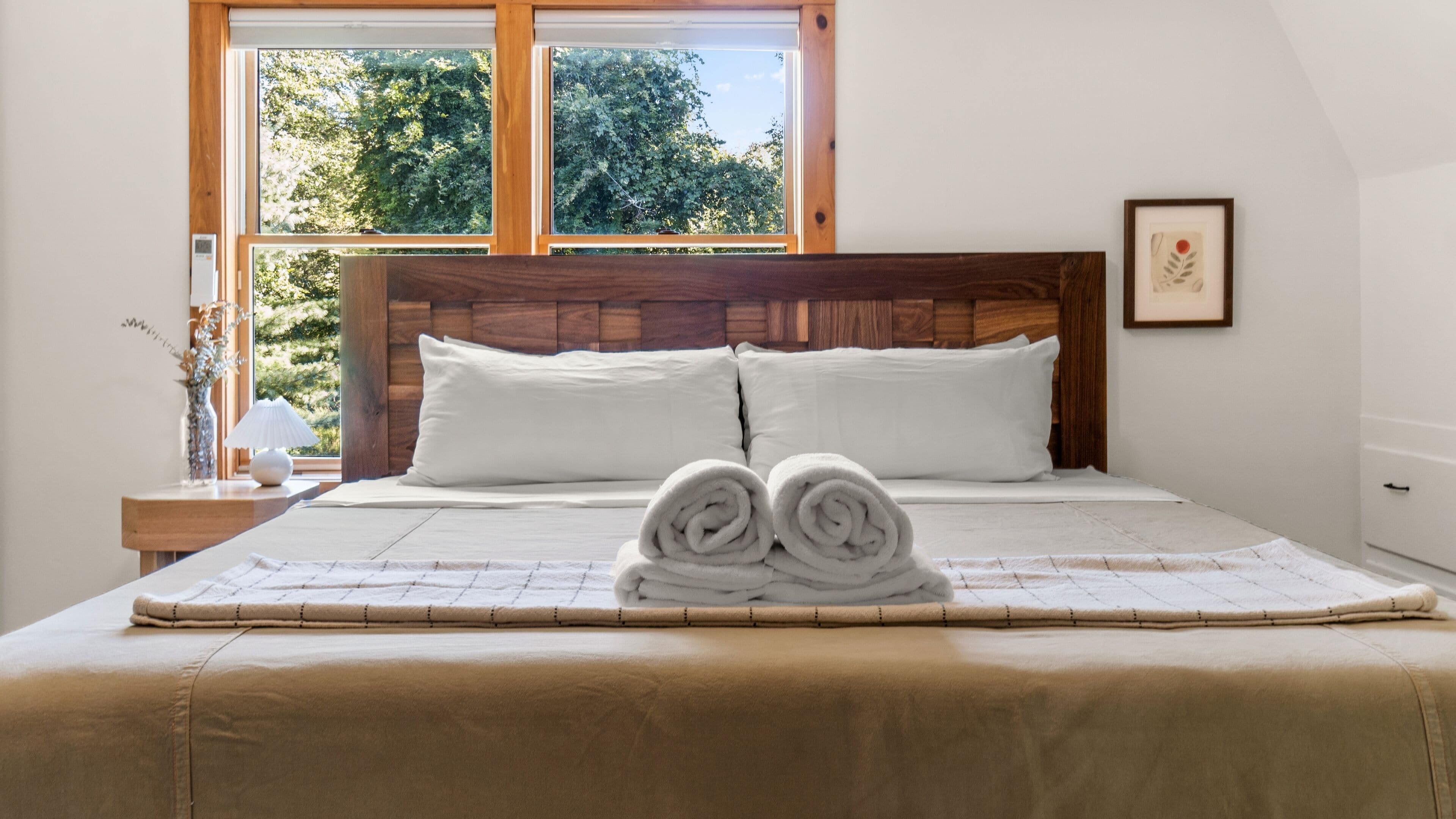 Een net opgemaakt bed met witte lakens en opgerolde handdoeken vooraan, een houten hoofdbord en een raam met uitzicht op bomen.