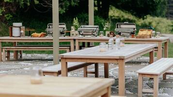 Outdoor wooden tables and benches arranged in a garden setting with greenery in the background.