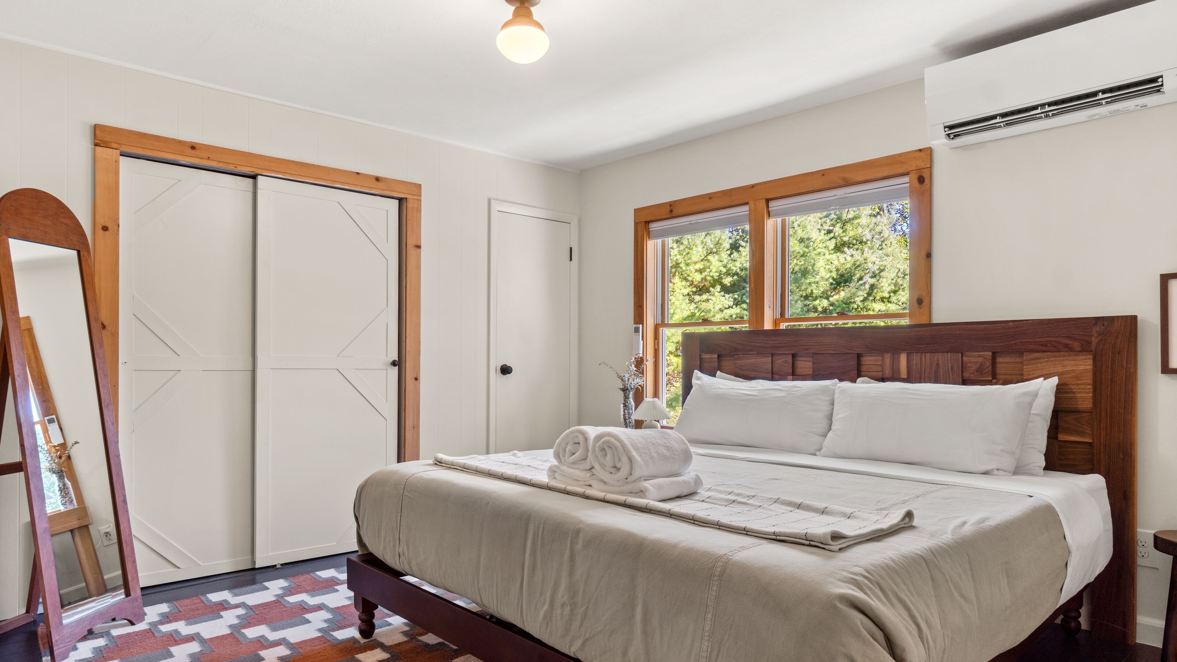 A bedroom with a bed, a wooden headboard, a window with a view of trees, and a door with a geometric design.