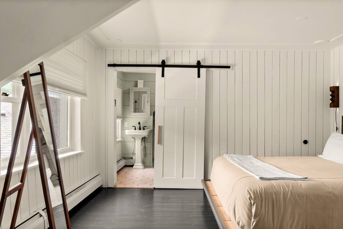 A bedroom with a bed, a wooden ladder, a window, and a door leading to a bathroom.