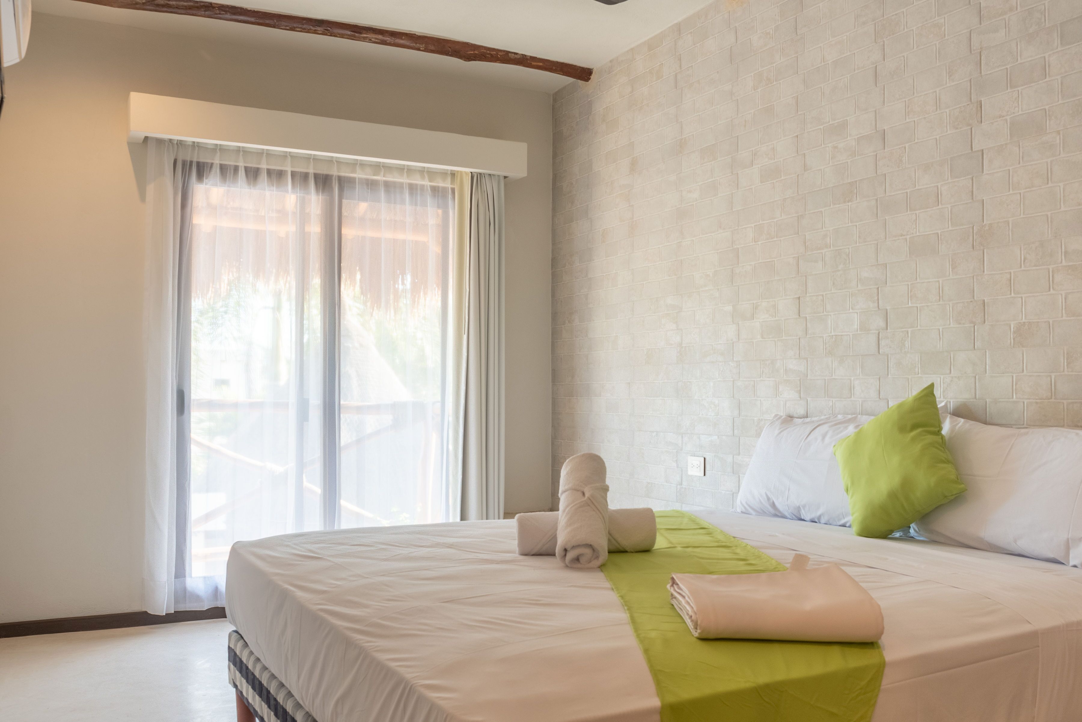 A hotel room with a bed, a green throw blanket, and a window with sheer curtains.