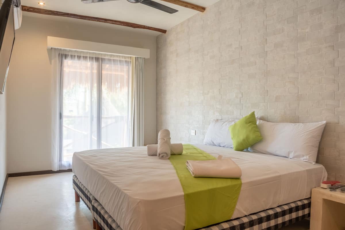 Ein Hotelzimmer mit einem großen Bett, einer grünen Tagesdecke, weißer Bettwäsche, einer karierten Bank, einem Nachttisch und einem Fenster mit durchsichtigen Vorhängen.