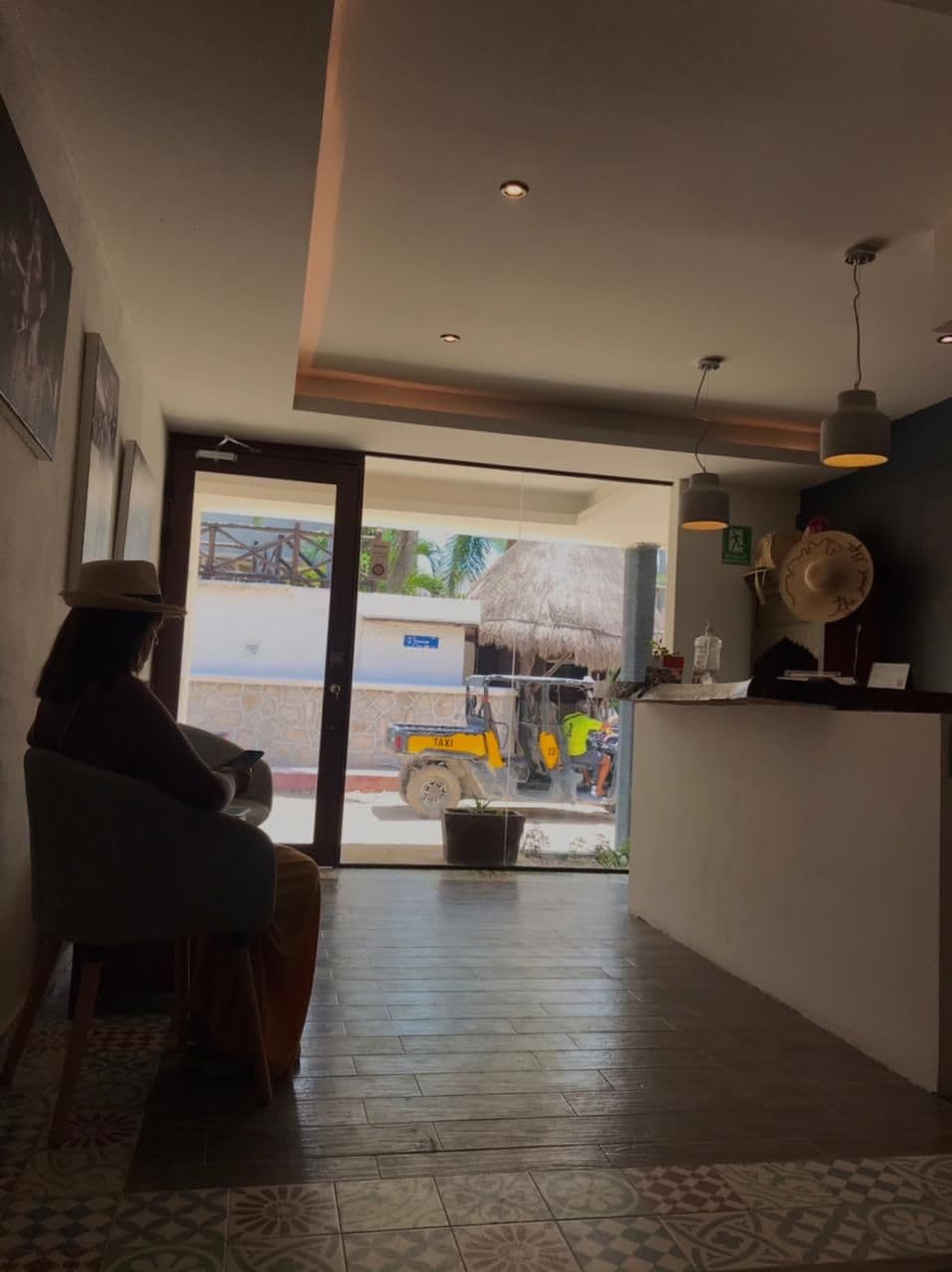 A hotel lobby with a person seated by a window, a reception desk, and a view of the street outside.