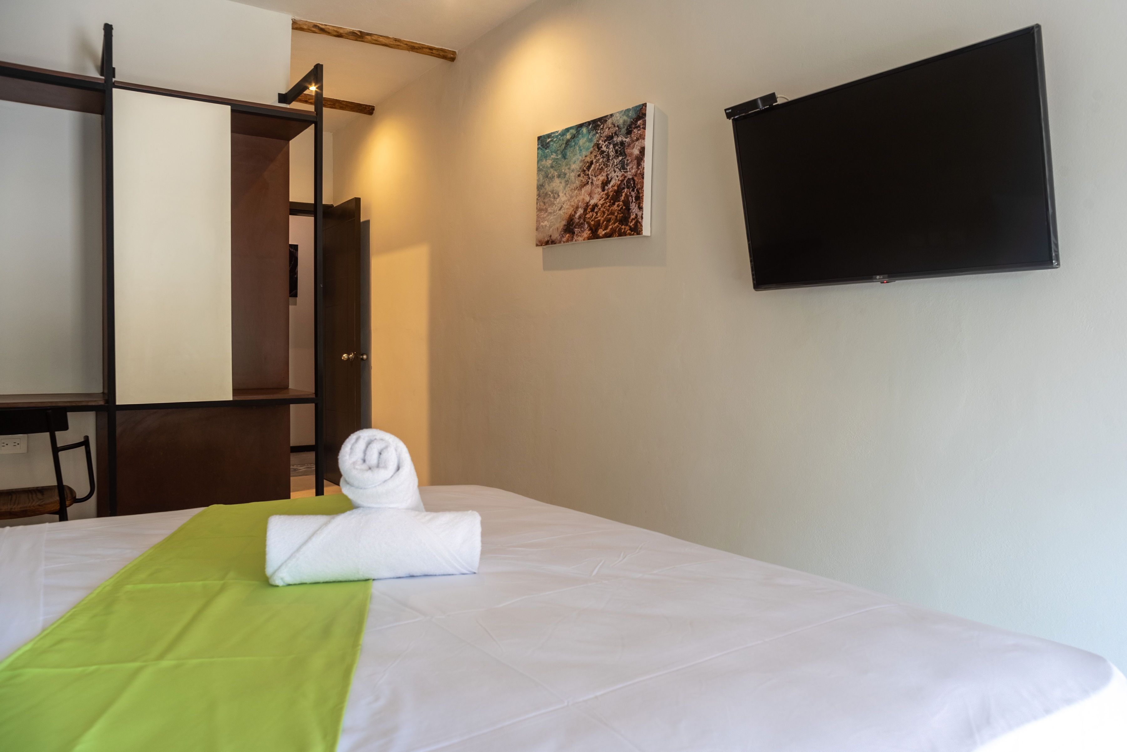A hotel room with a bed, a green bedspread, a rolled white towel, a painting, and a flat-screen TV.