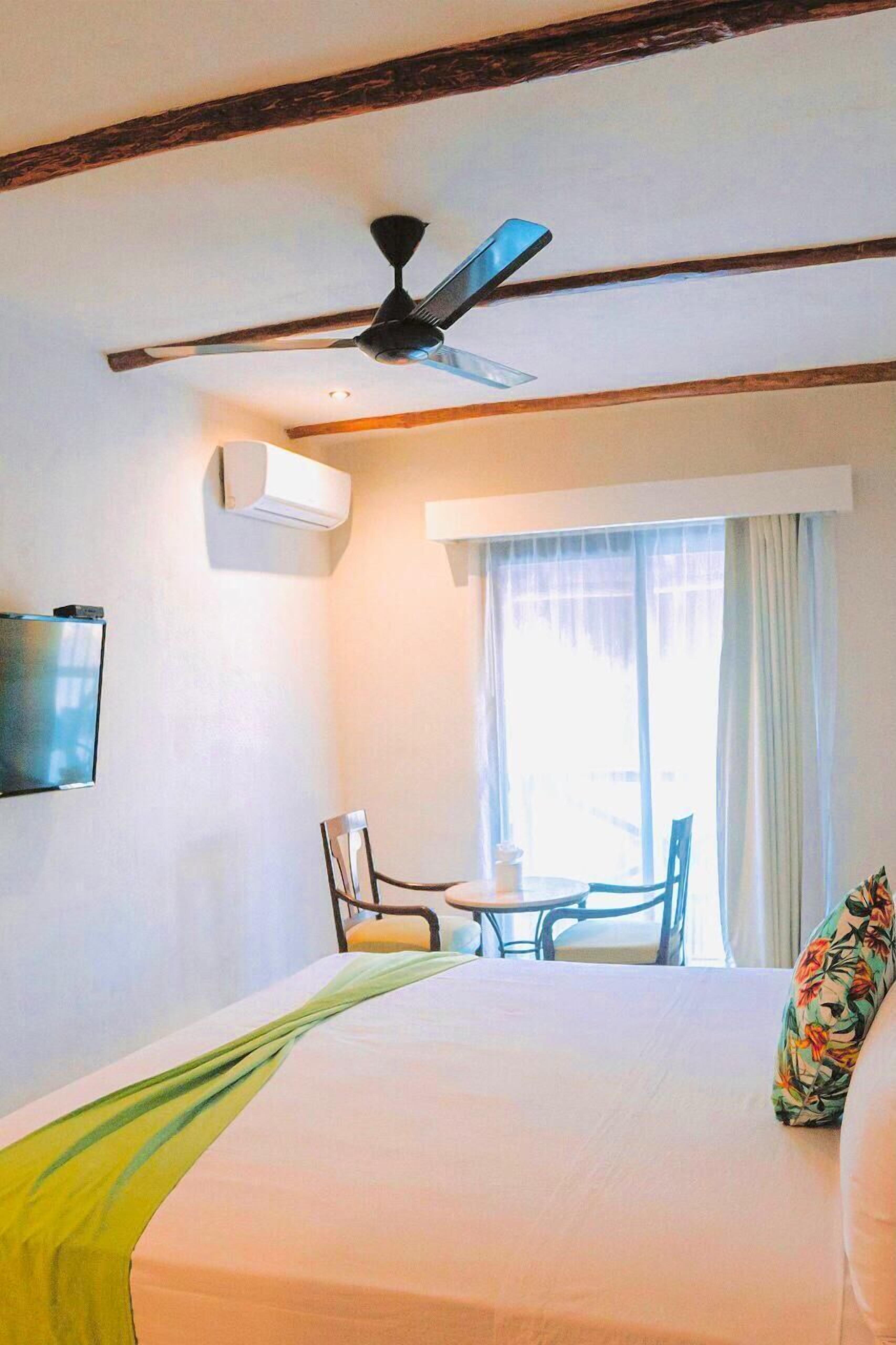 A hotel room with a bed, a ceiling fan, a TV mounted on the wall, a window with curtains, and a small table with chairs.