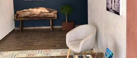 Een kamer met een houten bankje, een bloempot, een witte stoel en een tafel met tijdschriften en een ingelijste foto.