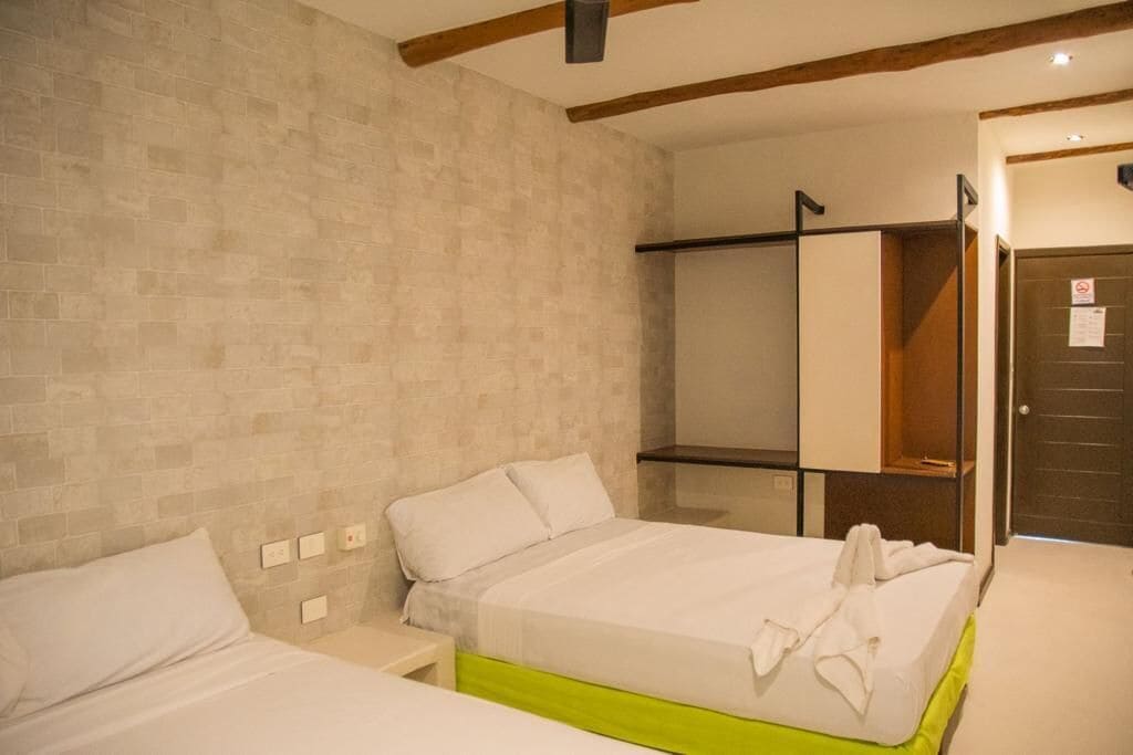 A hotel room with a brick-patterned wall, a bed with white bedding, a green bed skirt, a bedside table, and a wardrobe with a mirror.
