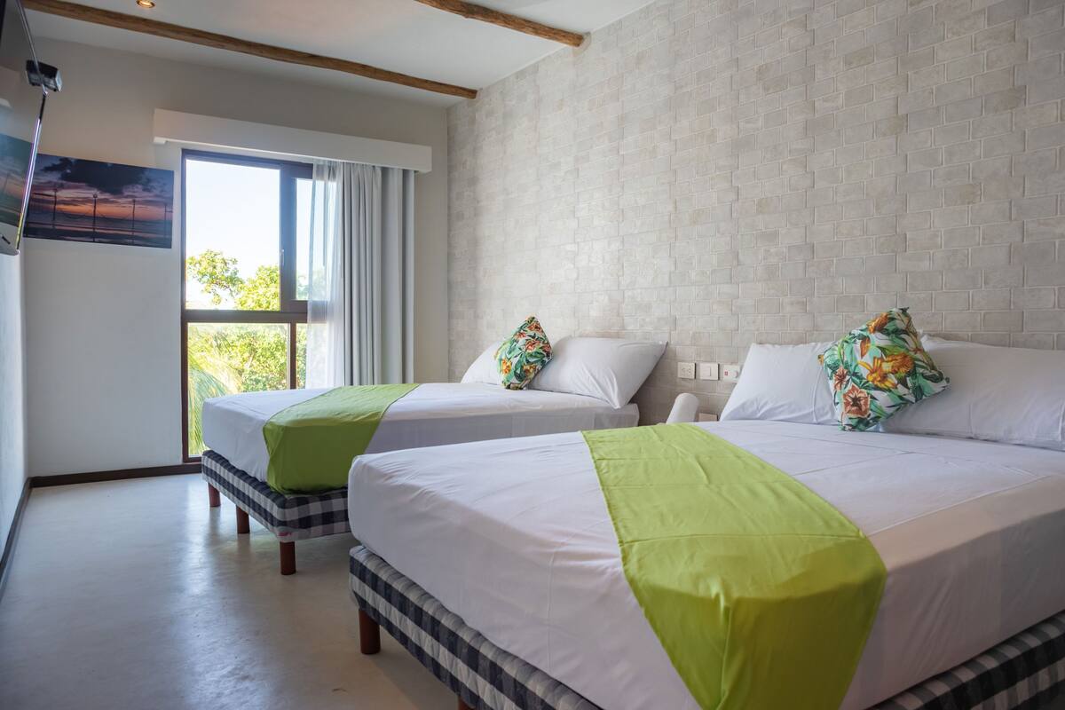 Ein Hotelzimmer mit zwei Betten, einem großen Fenster mit Vorhängen, einer Wand mit Ziegelmuster und einem Bild an der Wand.