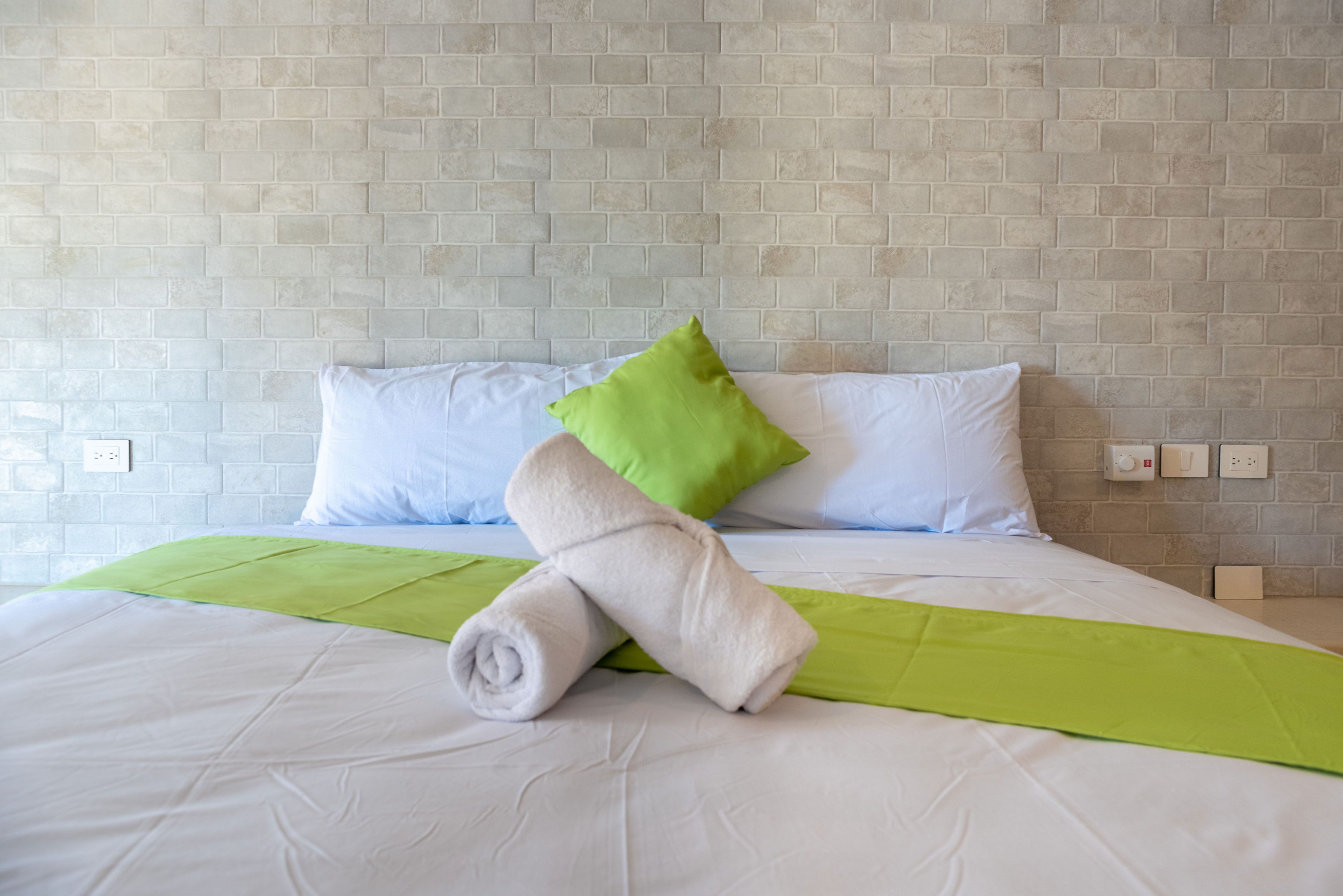 A neatly made bed with white linens and a green accent pillow, set against a light-colored brick wall.