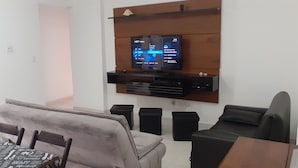 Ein modernes Wohnzimmer mit Flachbildfernseher an einer Holzwand, einem Essbereich mit Tisch und Stühlen sowie einem kleinen schwarzen Hocker.