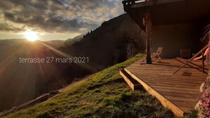 Una terraza de madera con sillas y una hamaca, con vistas a un paisaje montañoso al atardecer.