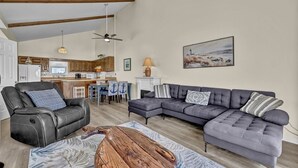 Ein Wohnzimmer mit Ecksofa, Ledersessel und Couchtisch. Im Hintergrund ist eine Küche mit Holzschränken und ein Essbereich mit blauen Stühlen zu sehen.