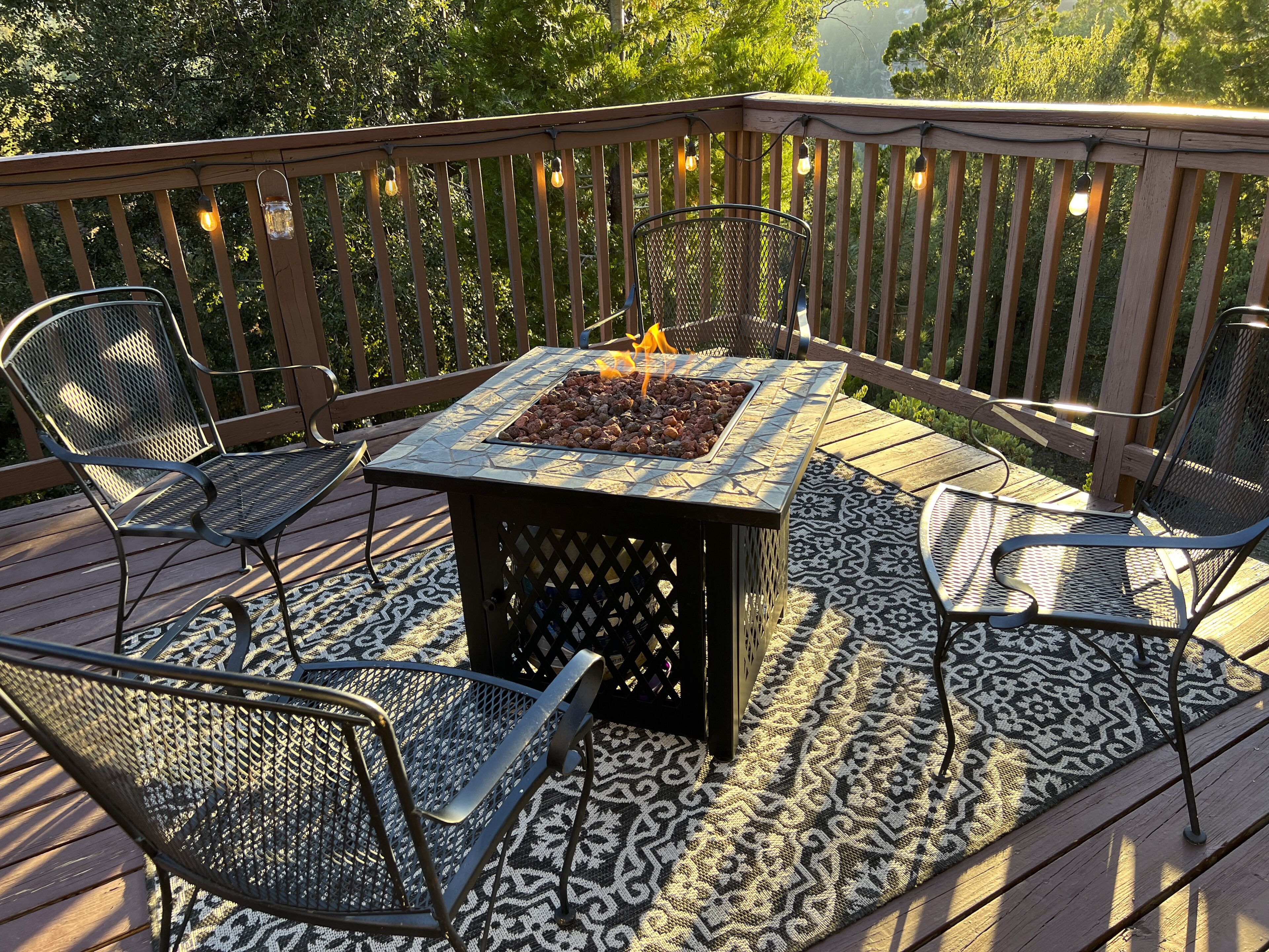 A deck with a fire pit table surrounded by metal chairs, a patterned rug, and a wooden railing.