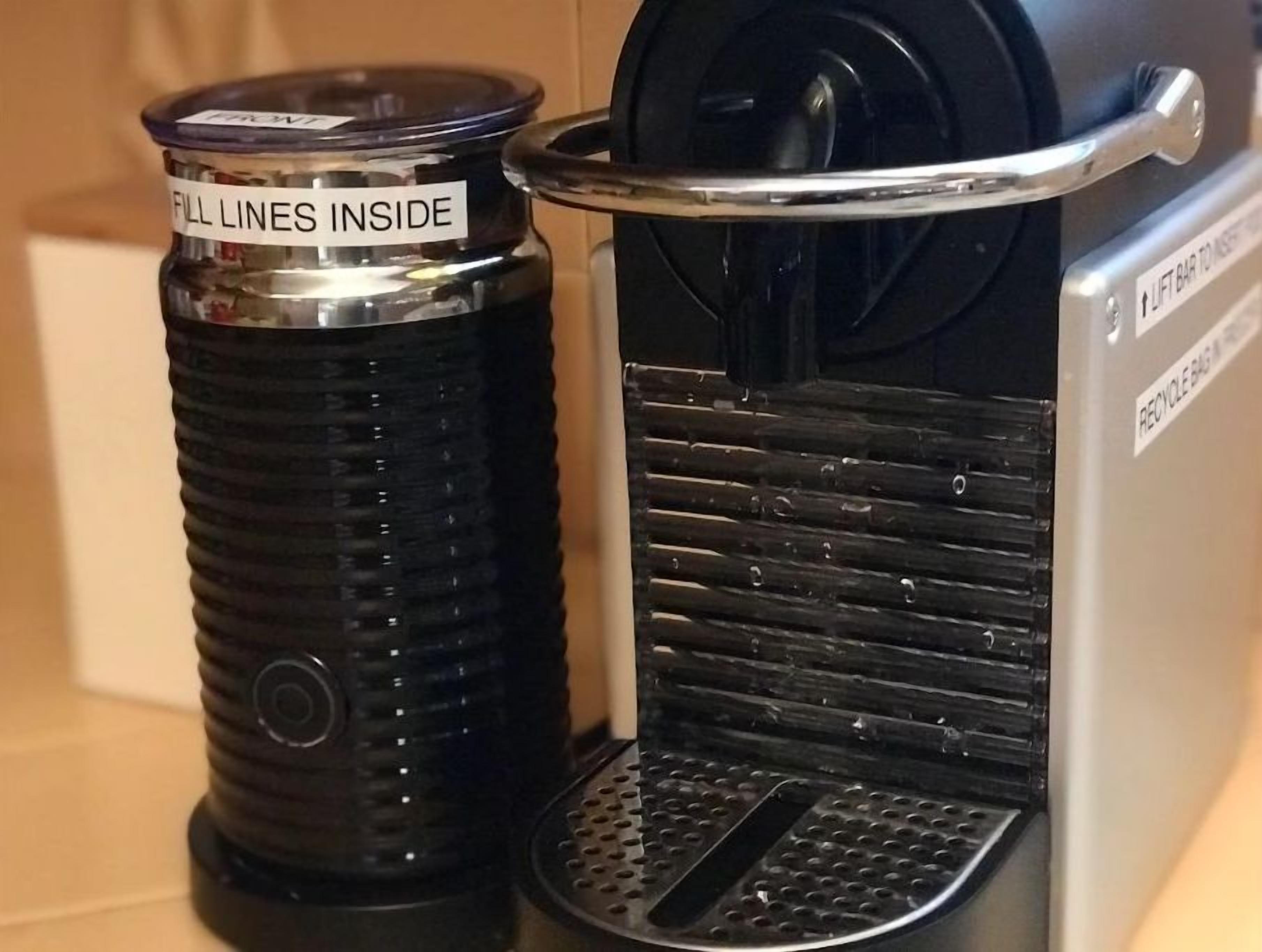 Una máquina de café con un rótulo que dice 'LLENAR DENTRO DE LAS LÍNEAS' y un contenedor de reciclaje con un cartel que indica 'BOLSA RECICLABLE EN SU INTERIOR'.