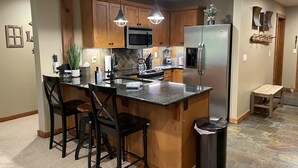 Una cocina moderna con armarios de madera, un refrigerador de acero inoxidable, un microondas y una encimera de granito. Se observan dos lámparas colgantes, un bote de basura y un taburete de madera pequeño.
