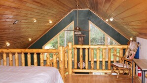 A room with a sloped ceiling, a bed, a rocking chair, and a view of trees through large windows. - Large Cabin with WiFi. Great Location: near Castle Rock + WI Dells (Friendship)