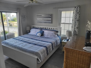 A bedroom with a bed, a nightstand, a lamp, a ceiling fan, and a view of the outdoors.