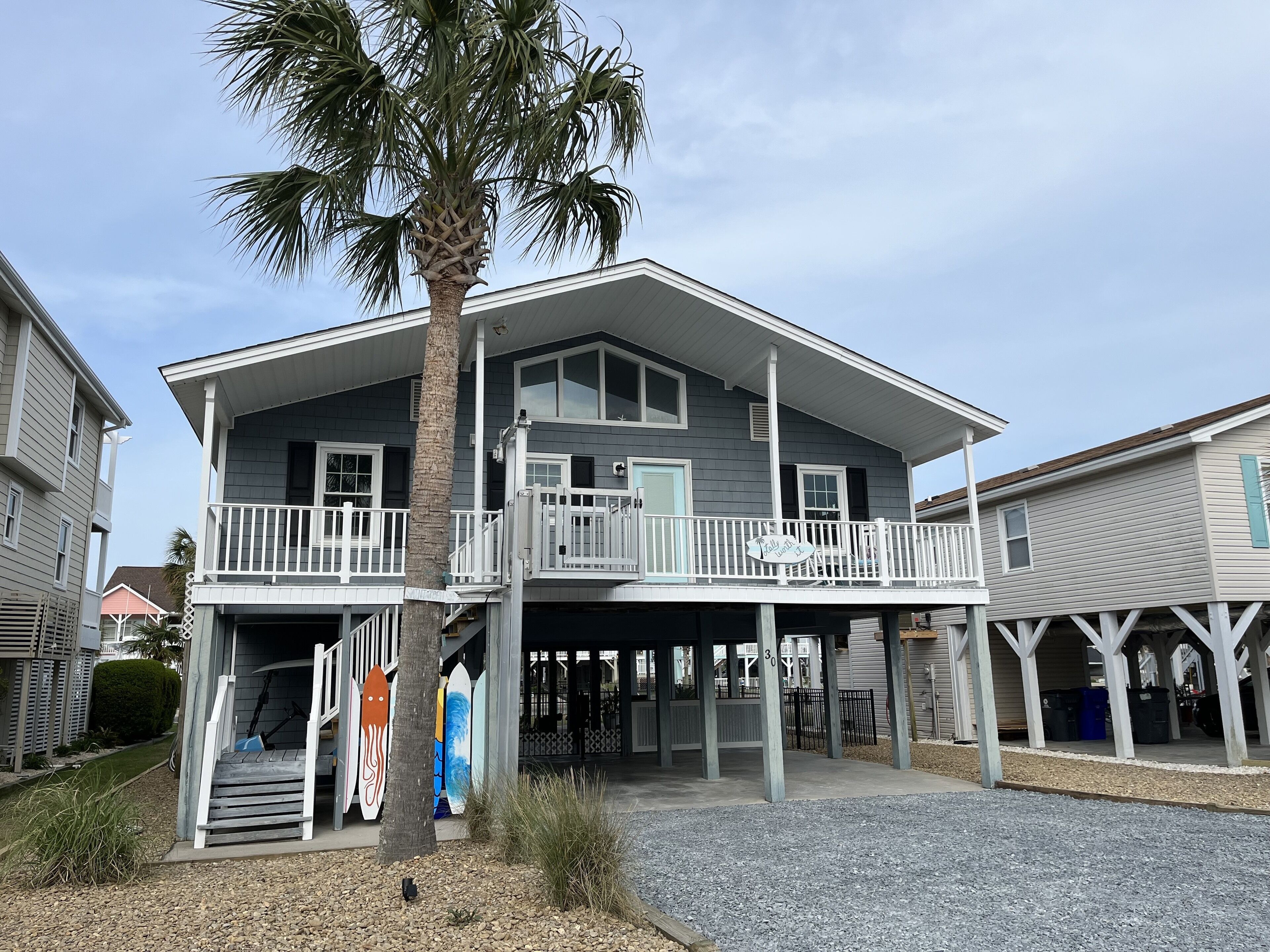 A two-story house with a balcony, a palm tree, and surfboards displayed outside.