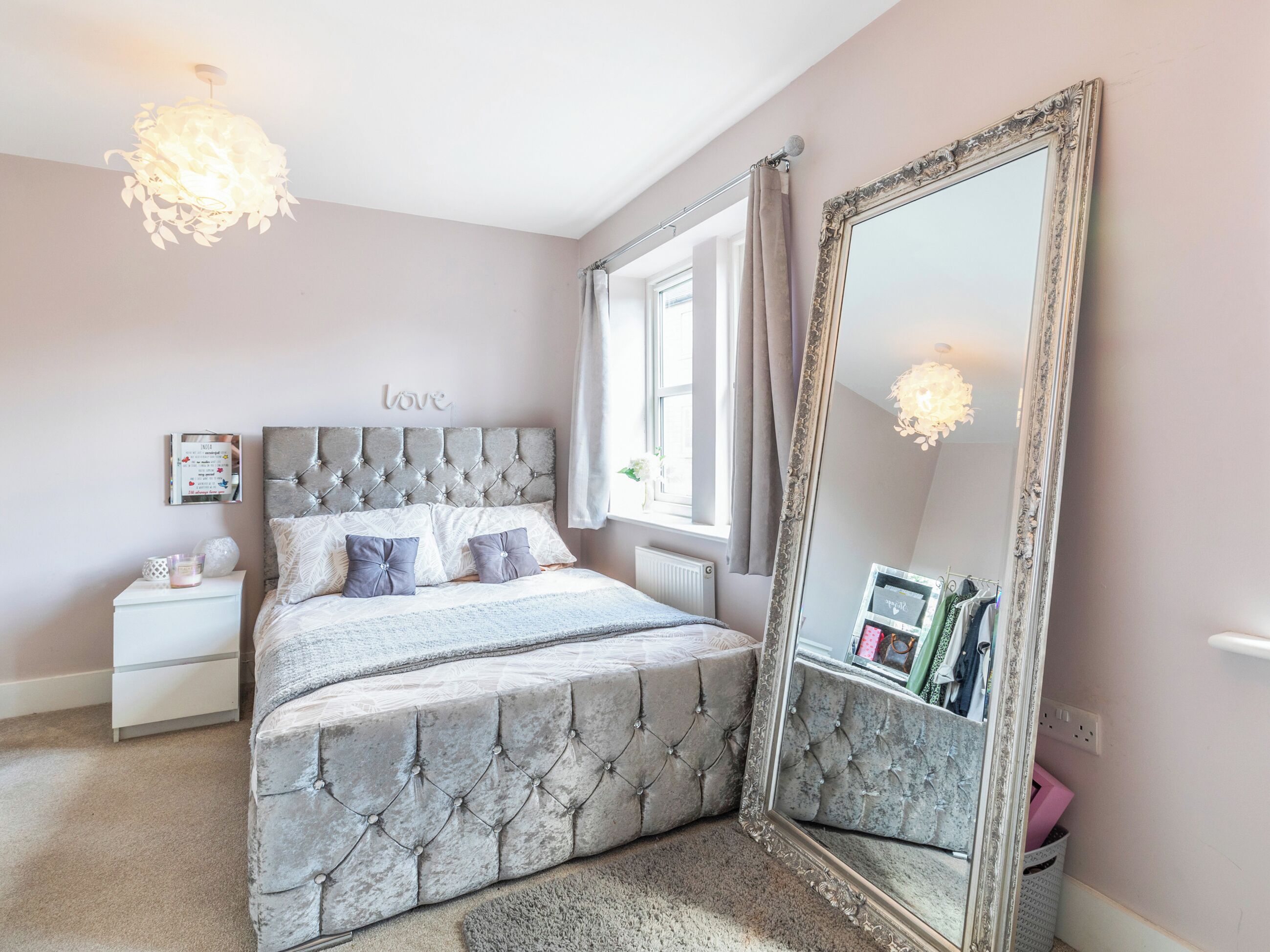 A bedroom with a tufted headboard, a large mirror, a bedside table, and a window with curtains.
