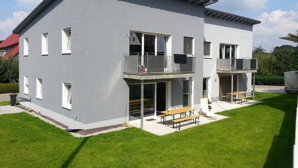 A modern two-story building with a flat roof, large windows, and a balcony.