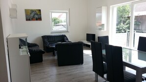 A modern living room with a black leather sofa, a glass dining table, and a TV. There is a window with a view of greenery outside.