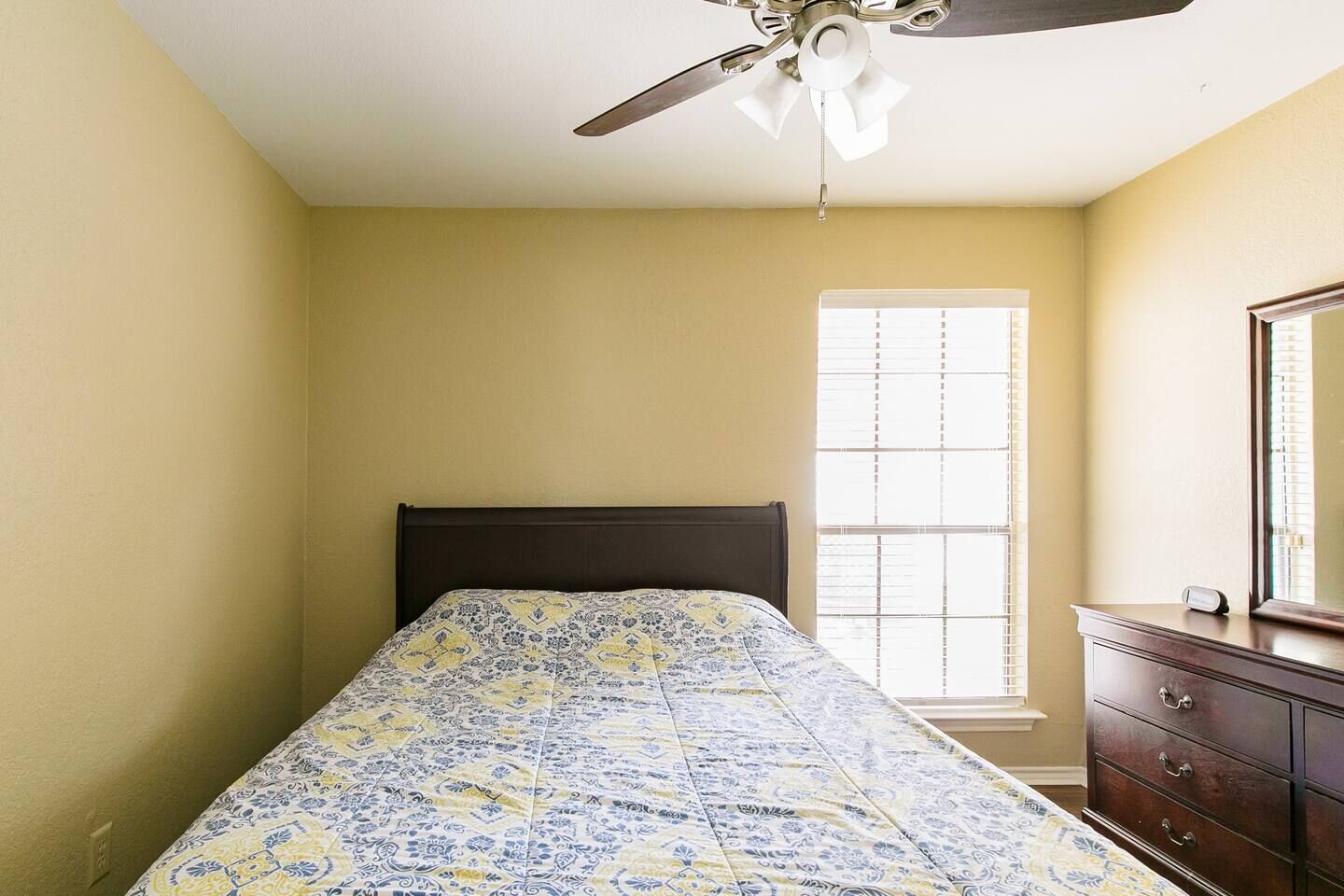 A bedroom with a bed, a ceiling fan, a window with blinds, and a dresser.