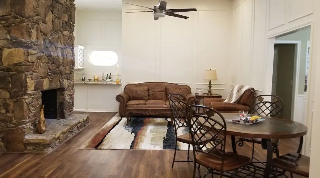 Un salon avec une cheminée en pierre, un canapé marron, une table à manger avec des chaises et un ventilateur de plafond.