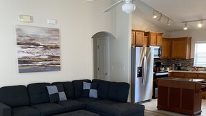 Una sala de estar moderna con un sofá modular, un ventilador de techo y una cocina con armarios de madera y un refrigerador.