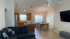 Una sala moderna con un sofá modular, un comedor con mesa y sillas, una cocina con armarios de madera y un refrigerador, y un ventilador de techo.