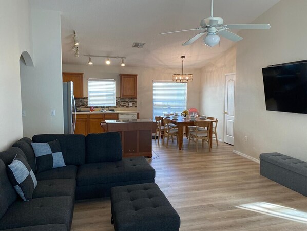A modern living room with a sectional sofa, a dining area with a table and chairs, a kitchen with wooden cabinets and a refrigerator, and a ceiling fan.