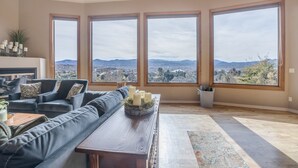A spacious living room with a large black leather sofa, a wooden coffee table with candles, and a view of a cityscape through large windows. - "The View in Prescott," a Custom Home With 3 Patios + Fire Pit, Near Downtown (Prescott)