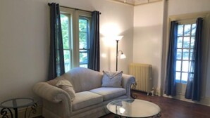 A living room with a sofa, a glass coffee table, and a floor lamp. There are windows with curtains and a radiator.