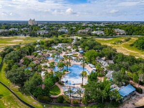 Luchtbeeld van een resort met een centraal gelegen zwembad, omringd door palmbomen en groen, met gebouwen op de achtergrond.