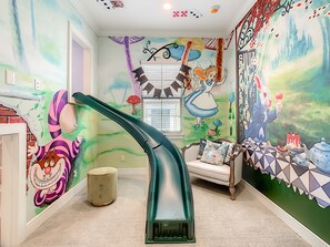 Una habitación con un tobogán verde, una silla blanca y un mural con un conejo y hongos.