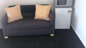 A dark grey sofa with two beige cushions, a small white refrigerator with a microwave on top, and a small potted plant.