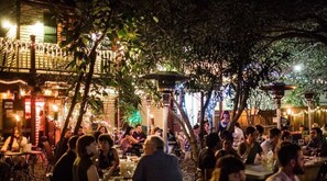 An outdoor dining area with tables and chairs, surrounded by trees and illuminated by string lights.