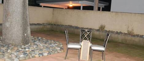 An outdoor patio area with a table and two chairs, surrounded by a stone border and a tree.