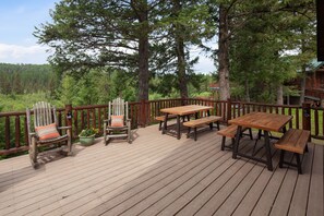 A wooden deck with a wooden table and benches, surrounded by a wooden railing, overlooking a forested area.