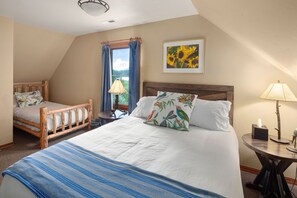A bedroom with a large bed, a smaller bed, a window with curtains, a painting, and a lamp on a side table.