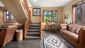 A spacious living area with a staircase, leather sofas, a desk, and large windows offering a view of the outdoors.