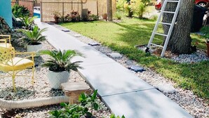 Un sendero conduce a una casa con un porche cubierto, plantas en macetas y una escalera apoyada en un árbol.