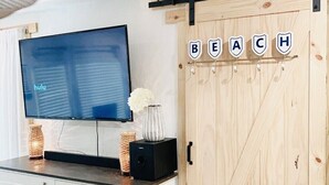 Una habitación con una televisión mostrando 'hulu', un tocador blanco con cajones, una puerta de madera con la palabra 'BEACH' escrita, y un pequeño florero con flores.