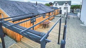 Een houten terras met een metalen hek, een overdekte zithoek en een gebouw met een donker dak.