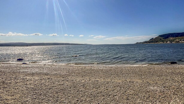 Een strand met kiezels, een heldere lucht en verafgelegen heuvels.