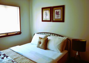 A neatly made bed with white linens and two pillows, a bedside lamp, and two framed botanical prints on the wall.