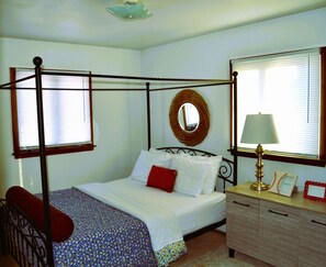 A bedroom with a four-poster bed, a bedside table with a lamp, a mirror, and a dresser.