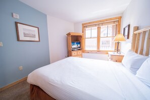 A bedroom with a bed, a wooden cabinet with a television, a window with blinds, and a lamp on a bedside table. - Buffalo Lodge 8352 (Keystone)