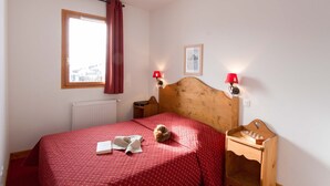 A bedroom with a wooden headboard, a red bedspread, a small wooden nightstand with a chessboard, two wall-mounted lamps, and a window with a curtain.