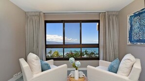Una habitación con dos sillones blancos orientados hacia una ventana grande que ofrece vista al océano. Una pequeña mesa de madera con un florero y dos tazas se encuentra frente a los sillones.