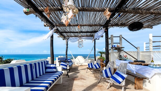 Eine Strandlounge mit blau-weißen Streifenmöbeln, einem Holztisch mit Getränken und Blick aufs Meer.